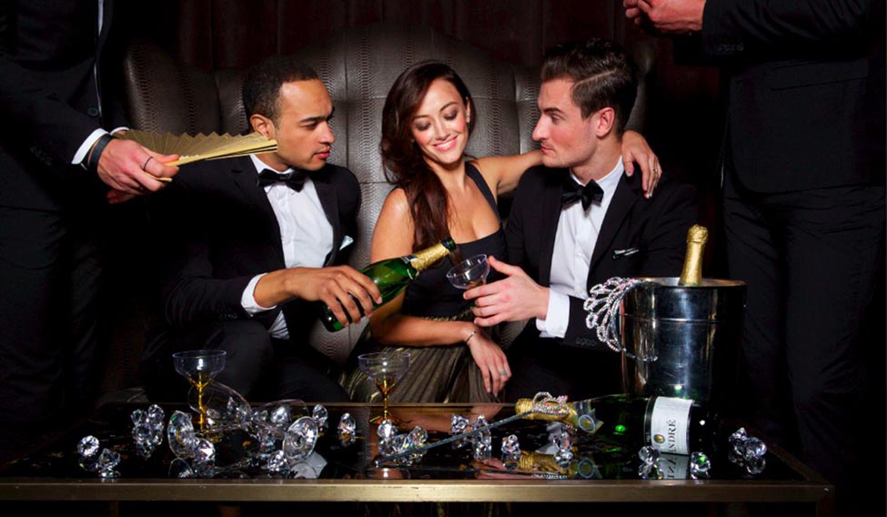 Orgazum911 VIP escort in black stunning dress with two men in tuxedos, champagne bottles, and waiter service—curated for upscale couple experiences